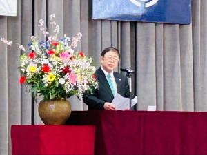 演台横に白、ピンク、黄色、赤と色鮮やかな花が活けられた花瓶が飾られマイクの前で千徳小学校卒業証書授与式の挨拶をしている市長の写真