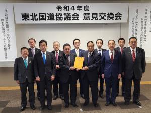 「令和4年度東北国道協議会意見交換会」と書かれた横断幕が掲げられた会場で、国土交通省の長谷川朋弘国道・技術課長と谷藤裕明会長が要望書を一緒に持ち、市長を含め9名の県内首長の方々と横一列に並んでいる写真