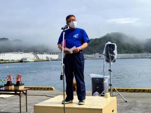 港に置かれた木製の台の上に立ち、スタンドマイクの前で話をしている様子の市長の写真