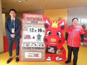 赤いユニフォーム市長とスーツ姿の岩手ビッグブルズの社長が試合日程の看板と紙でできた岩手ビッグブルズのマスコットをはさむように立って撮影した写真
