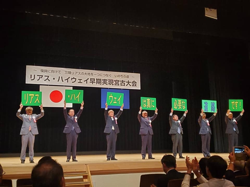 「リアス・ハイウェイ早期実現宮古大会」と書かれた横断幕が吊るされた舞台上で、3文字ずつ文字が書かれた緑色のパネルを頭上に掲げて横一列に並ぶ参加者の方々7名の写真