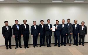白い壁をバックに、中央に文部科学省研究振興局長の森晃憲氏と市長会の代表が一緒に要望書を持って中央に立ち、市長を含む9名の関係者が横一列に並んでいる写真