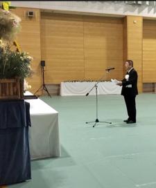 市長が黒のスーツに白の胸章をつけ、献花台の前のスタンドマイクで式辞用紙を読み上げている写真