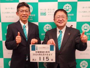 宮古市のバックパネルの前で「全線開通5周年 2024年6月9日まであと115日」と書かれた用紙を田村省二所長と市長が持ちながら親指だけあげてグーのポーズをしている写真