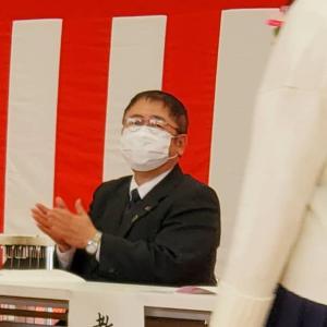 紅白幕の前に置かれたテーブルと椅子に腰かけ拍手をしている様子のマスクを着けた市長の写真