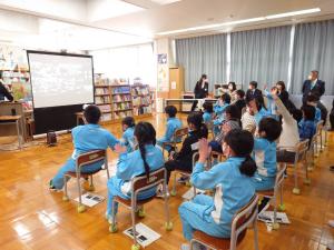 上下水色のジャージ姿の川井小学校の児童たちとスクリーンに映った青山学院初等部6年生とオンライン学校交流をしているのを、市長、宮古市に滞在の青山学院大学の学生たちが見守っている写真