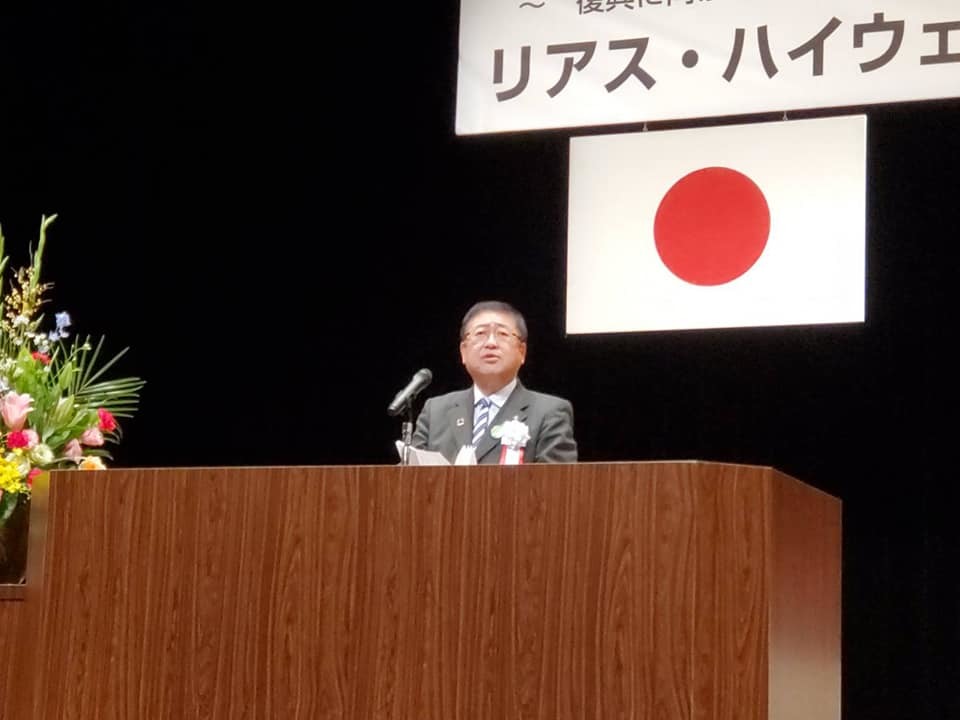 日本国旗が吊るされた壇上に置かれた演台の前で話をする様子の市長