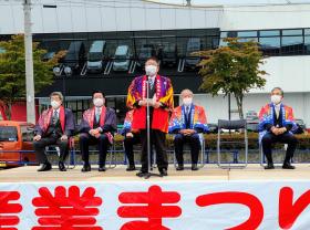 法被を着た来賓の方々が座っているまつり会場のステージ上で、法被を着た市長がマイクの前であいさつをしている写真