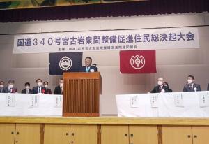 「国道340号宮古岩泉間整備促進住民総決起大会」と書かれた横断幕が舞台上に掛けられ、舞台の両側には白いテーブルクロスを引いた机の前に座る関係者の方々、中央に置かれた演台の前で話す様子の市長が写った写真