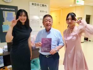 ピースサインをしている黒のワンピースを着た女性とピンクのワンピースを着た女性の間にパンフレットを持った市長が立っている写真