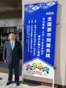 全国都市問題会議の立て看板の横に立つ市長の写真