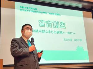 「宮古創生」という文字が映されたスクリーンを背に、右手でマイクを持ち話している様子の市長の写真