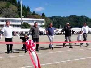 大人の男性5名の間に子供2名が入り、お互い手を繋いで、横一列に並んでグランドを歩いている写真
