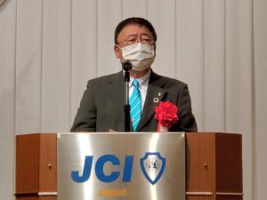 JCIのロゴマークが表示された檀上で、胸章リボンを付けた市長が挨拶を行っている写真