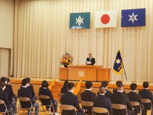 舞台上の演台の前で話す様子の市長と、舞台の下で椅子に座って演説を聞いている様子の学生たちの写真