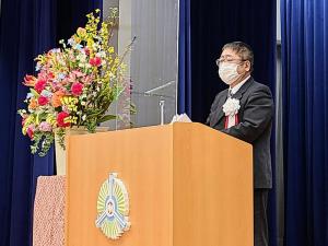 色とりどりの花が活けられた大きな花瓶が隣に置かれた演台の前で、白い胸章を着けて話をする様子のマスクを着けた市長の写真