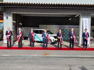 導入した電気自動車を背景にレッドカーペットの上に立った市長の他6人で、紅白のテープカットした写真