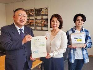 ラーメン店「野菜たっぷりタンメンの店・大久保」の店内で、女性と市長が一緒に証を持ち、義援金と書かれた箱を持った右側の女性と横一列に並んでいる写真