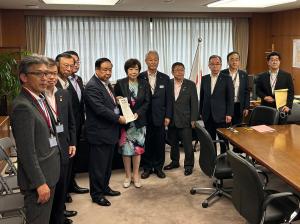国旗が設置された室内で、中央に経済産業省の太田房江副大臣と市長会の代表が一緒に要望書を持って中央に立ち、市長を含む9名の関係者が横一列に並んでいる写真