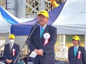 スーツ姿に黄色のキャップを被った市長がマイクの前に立ち挨拶を行っている様子の写真