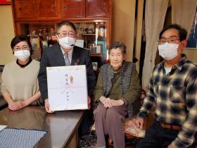 鳥居キヨヱさんのご自宅でお祝いの記念品を持った市長と鳥居キヨヱさんとご家族2人で居間に座って撮影した写真