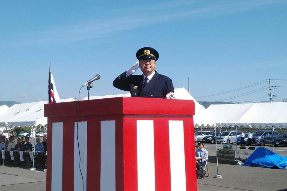 紅白に塗られた演台の前に立ち、制帽と制服を身に着け白い手袋を着けた右手で敬礼をしている市長の写真
