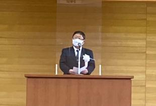 アクリル板が置かれた演台の前で胸章をつけた市長があいさつを述べている写真