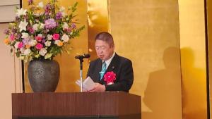 金屛風の前で、花が飾られた演台で市長が赤の胸章をつけ、式辞用紙を開いて読んでいる写真