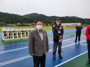 青いタータンが敷かれた陸上競技場のトラックの上で、首を斜め前に向けて立っている市長と関係者の方々3名が写った写真
