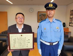 賞状の額縁を両手で持って正面に向ける市長と、制服を着て制帽を被った警官が並んで写っている写真