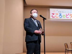 舞台上に置かれたスタンドマイクの前で、両手に資料を持ち話している様子のマスクを着けた市長の写真