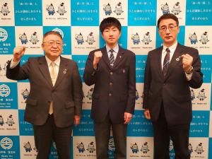 バックパネルの前で男子学生を中心に市長と関係者の男性が横一列に並び、全員が片手の握りこぶしを胸の横に掲げている写真