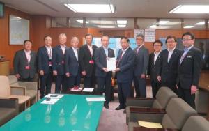 応接椅子やテーブルが設置された室内で、中央に国土交通省の吉岡幹夫技監と市長会の代表が一緒に要望書を持って中央に立ち、市長を含む9名の関係者が横一列に並んでいる写真