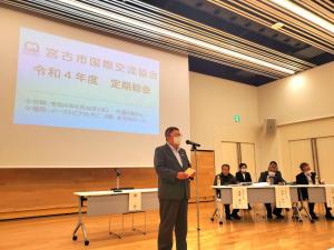 「宮古市国際交流協会 令和4年度定期総会」と書かれた画面が映されたスクリーンが設置された会場の中心で、スタンドマイクの前で話をする様子の市長と斜め後方で長机の前に座る関係者たちを写した写真