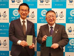 バックパネルの前で長方形の形をした薄黄色の用紙を持つ名取市の山田司郎市長と、緑色の用紙を持つ市長が並んでいる写真