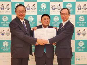 バックパネルの前で締結書を両手で持ち広げる市長と、市長の両側に立ち片手を伸ばして市長の前で握手し、もう片方の手は市長の肩に乗せている男性2人を写した写真