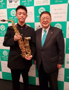 宮古市のバックパネルをバックにサクソフォーンを持った大村光介さんと市長が並んで記念撮影をしている写真