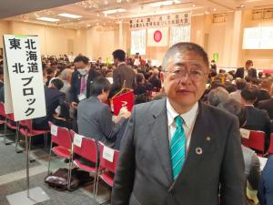 椅子に座っている大勢がいる会場を背に、参加者の後ろで立つ市長を正面から写した写真