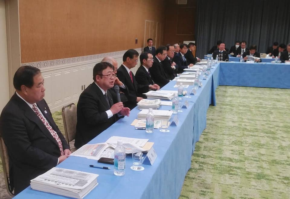水色のテーブルクロスが掛けられた長机の席に参加者が座り、手前から二番目に座った市長が右手にマイクを持ち話をしている様子の写真