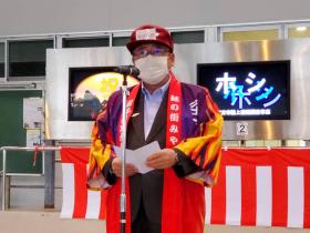赤い帽子を被り、法被を着た市長が紅白の幕が張られた台に置かれた2台のモニターを 背景にあいさつをしている写真