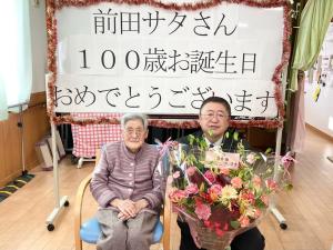 「前田サタさん100歳お誕生日おめでとうございます」と書かれた紙が貼られたホワイトボードの前で、椅子にすわる前田サタさんと花束を持った市長の写真