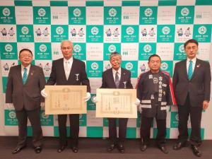 バックパネルの前で左から市長、白い手袋をして賞状を持った男性が2人、法被を着ている男性とスーツ姿の男性が横一列に並んでいる記念写真