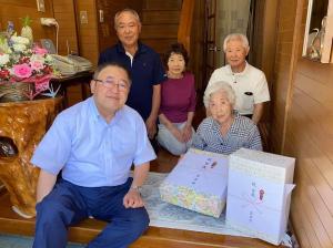 玄関先に座る市長、贈呈品の後に座る満100歳を迎えた菅原ノリさんと、その後方に座るご家族の方々3名を写した写真