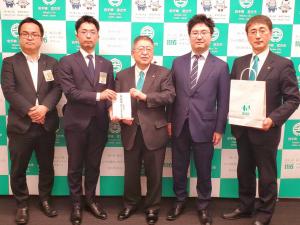 宮古市のバックパネルをバックにスーツ姿の男性5名が1列に並び、左から2番目の男性と中央の市長が義援金を持ち、1番右の男性が紙袋を持ちながら記念撮影をしている写真