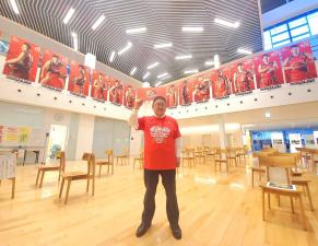 市長が赤のTシャツを着て、岩手ビッグブルズの選手のポスターが一面に貼ってある会場で右手でガッツポーズをしている写真