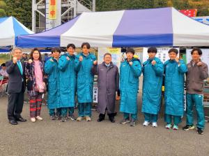 青と白のストライプ柄のテントの前で、市長を中央に青山学院大学陸上競技部の7名の選手と2名の関係者が横一列に並びガッツポーズをしている記念写真