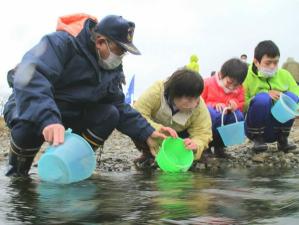 津軽石川の河川敷で、放流する前のさけ稚魚が入った水色のバケツを持った3名の子供たちが並んで座り、左側に座っている市長が子供たちの放流を手伝っている様子の写真