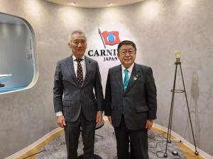 灰色の壁に旗の様なマークが描かれた室内の一角で、カーニバル・ジャパン代表の堀川社長と市長が並んでいる記念写真