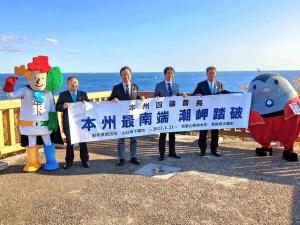 海が見える青空の下、本州最南端潮岬踏破と書かれた横断幕を市長含め男性4名が手に持ち、本州四端協議会マスコットキャラクターのよんたんと串本町マスコットキャラクターのまぐトルが両端に立っている写真