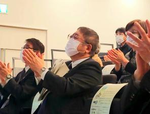 階段状に椅子が配置された会場で、椅子に座り資料を抱えて拍手をする様子の市長と参加者の方々を斜め前から写した写真